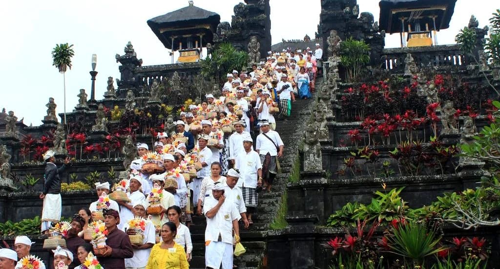 Devotees in white robes ascend stone steps at Besakih Mother Temple, surrounded by statues and vibrant flowers.